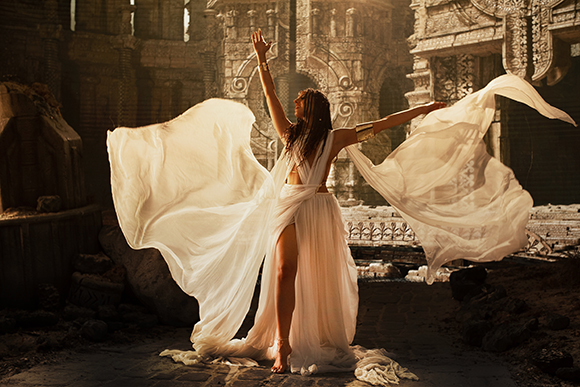 A woman in a long, flowing white dress poses with arms outstretched against ancient stone ruins.