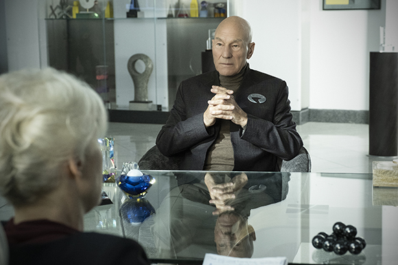 A bald man in a dark blazer sits at a glass table with his hands clasped, facing a person with blonde hair.