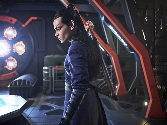 A person with pointed ears and long dark hair wears blue robes and holds a staff in a futuristic setting.