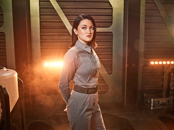 A woman with brown hair wearing a grey jumpsuit stands in a dimly lit, industrial sci-fi setting.