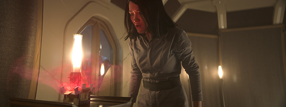 A woman in a grey long-sleeved shirt leans toward a bright, glowing light in a dark room.
