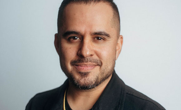 A man with short dark hair and a salt-and-pepper beard smiling against a light gray background.