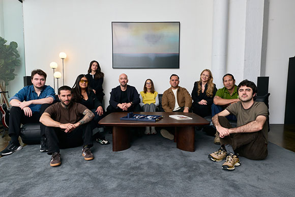 A diverse group of eleven people posing together in a bright, modern room with a large painting on the wall.