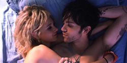 A blonde woman and a dark-haired man with tattoos lie close together on blue bedding.