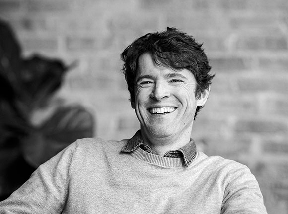 A smiling man with dark wavy hair wearing a sweater over a collared shirt.