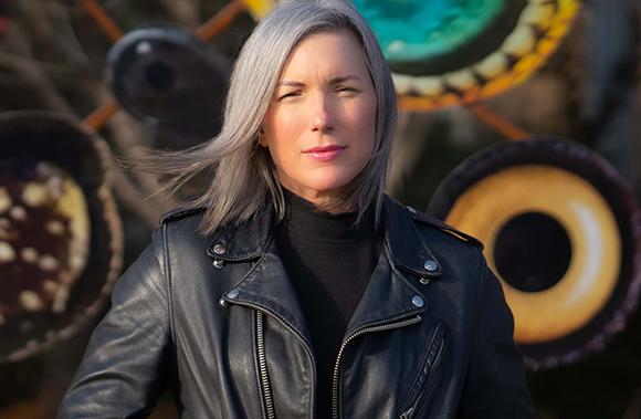 A woman with silver hair and a black leather jacket stands before colorful circular art pieces.