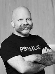 Black and white portrait of a bald man with a large mustache and arms crossed, showing a tattoo.