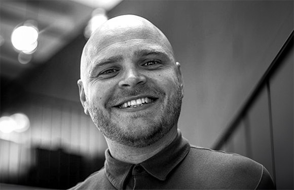 Black and white close-up portrait of a smiling bald man wearing a polo shirt.