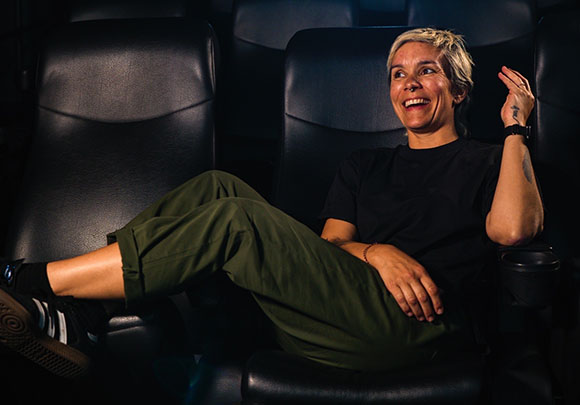 A blonde woman in a black t-shirt and green pants smiles while sitting in a dark cinema seat.