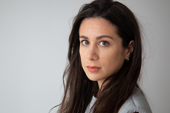 A woman with long dark hair and brown eyes looks directly at the camera against a plain light background.