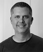 Black and white headshot of a smiling man with short hair and light facial stubble.
