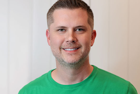 A smiling man with short brown hair and light facial hair wearing a green t-shirt against a white background.