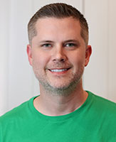 A smiling man with short brown hair wearing a green crewneck t-shirt against a white background.