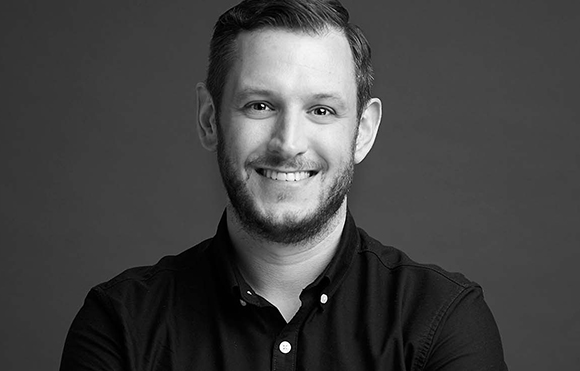 Black and white headshot of a smiling man with a beard wearing a dark button-down shirt.