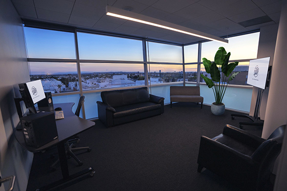 Office lounge with a black leather sofa, desk, and large windows showing a city skyline at sunset.
