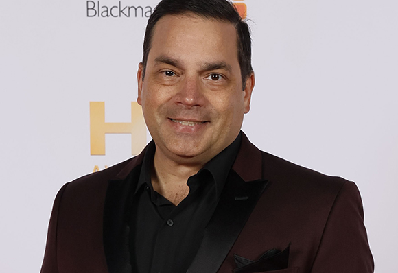A man with short dark hair smiling while wearing a black shirt and a maroon blazer.