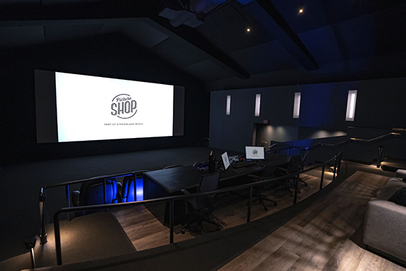 A dark screening room featuring a large white projection screen and a desk with computer monitors.