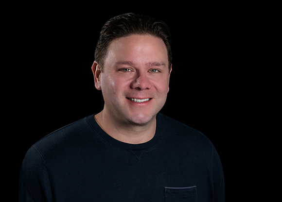 A smiling man with short dark hair wearing a black long-sleeved shirt against a black background.
