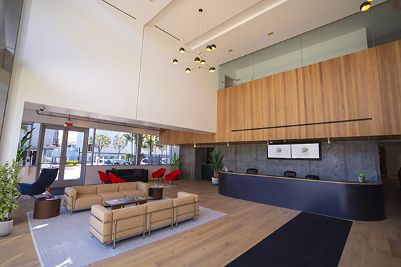 A spacious, modern office lobby with a long dark reception desk, wooden walls, and a tan leather seating area.