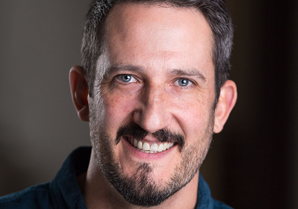 A close-up portrait of a smiling man with short brown hair and a dark beard.