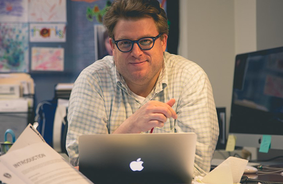 A man with glasses and a light checkered shirt sits at a desk behind an open laptop.