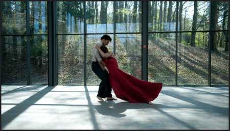 A man and a woman in formal attire dance together in a large room with floor-to-ceiling windows.