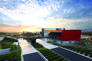 Aerial view of a large modern building with a red curved wall under a bright sunset sky.