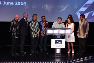 A group of men and a woman stand on a stage behind a large film strip prop with a logo.