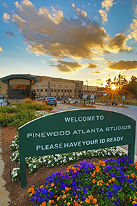 A green sign reads 'Welcome to Pinewood Atlanta Studios' in front of a large building under a sunset sky.