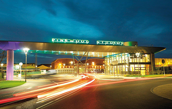 The glowing Pinewood Studios sign atop a modern building entrance with light trails from passing cars.