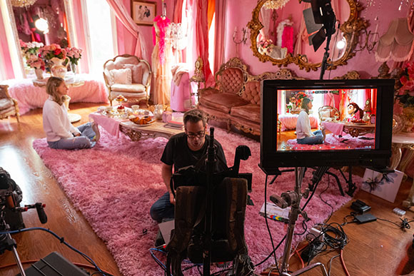 A camera monitor shows a woman sitting in a pink room while a crew films in a studio set.