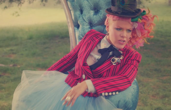 A person with pink hair wearing a red striped blazer and blue tulle skirt sits in a tufted blue chair.