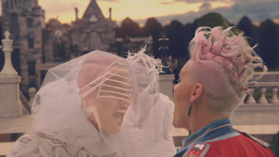 Two people with pink hair and avant-garde white and red costumes face each other near a stone building.