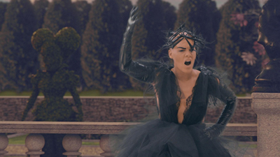 A person in a black feathered gown and headpiece poses in a formal garden with topiary.