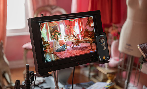 A film monitor shows a woman in a pink room, with a smartphone beside it displaying a digital character.