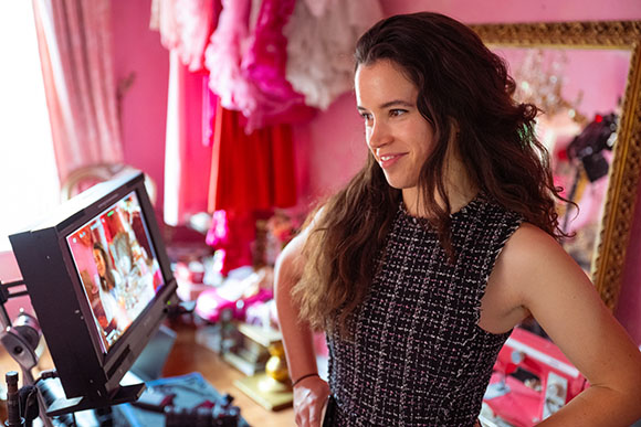 A woman with long brown hair smiles while looking at a video monitor in a pink room.