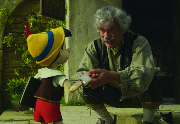 A wooden puppet in a yellow hat holds hands with a gray-haired man crouching in front of a stone building.