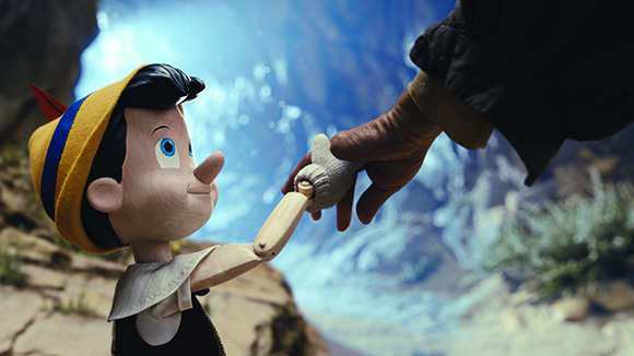 A small wooden puppet boy reaches his arm up toward a large human hand against a blurred mountain background.