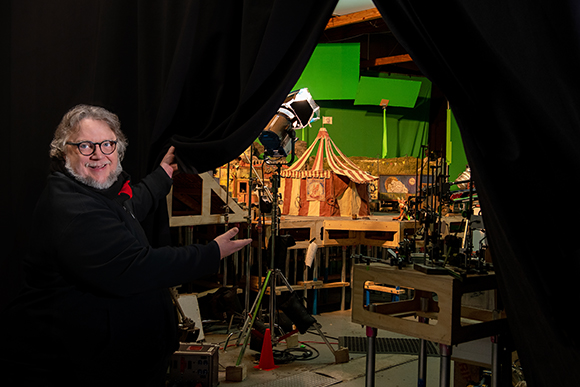 A man with glasses and a beard gestures toward a miniature circus set behind black curtains.