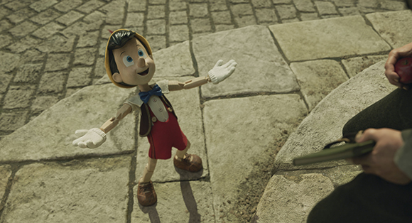 A wooden puppet boy with large eyes and a yellow hat dances on a stone street.