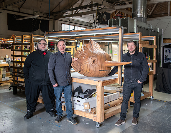 Three men pose behind a large, carved wooden fish sculpture on a workbench in a workshop.