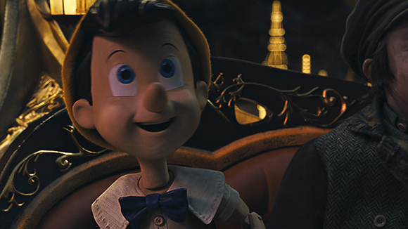 A smiling wooden puppet with large blue eyes and a blue bowtie sitting in an ornate carriage.