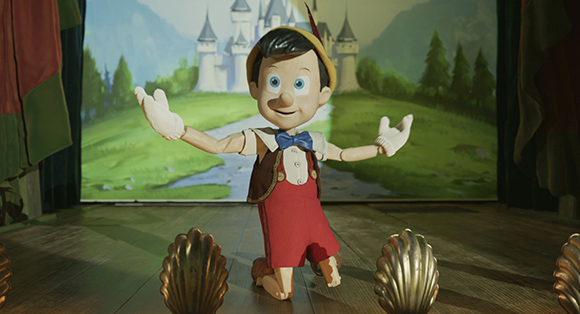 A wooden puppet with large eyes and a yellow hat gestures with open arms on a stage with a castle backdrop.