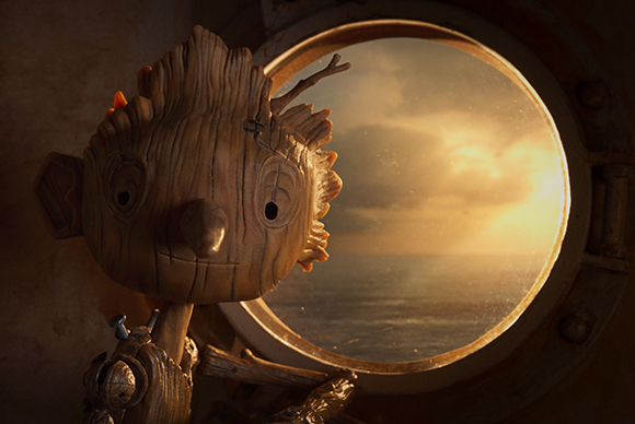 A small wooden creature gazes through a circular porthole at a golden sunset over the sea.