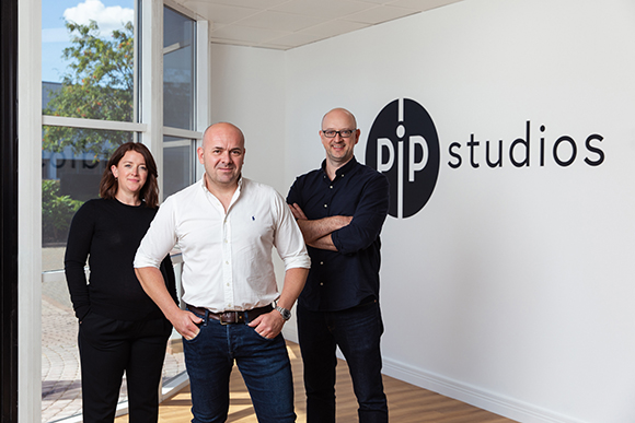 Three people stand together in front of a white wall featuring a black circular logo and text.