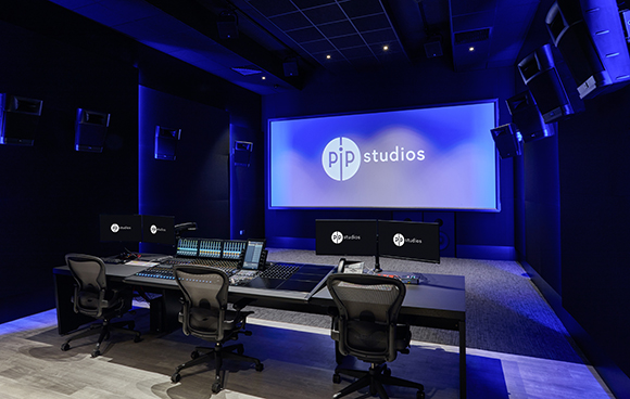A dark audio production studio featuring a large screen with 'pip studios' logo and a mixing console.