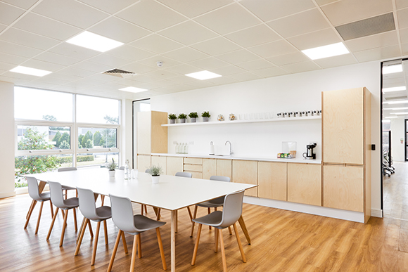 A bright office breakroom with a long white table, grey chairs, and light wood cabinetry.