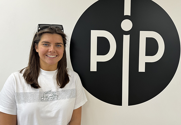 A smiling woman in a white t-shirt stands in front of a large black circular logo containing the letters 'pip'.