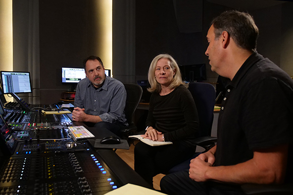 Three people sit around a large audio mixing console in a dimly lit recording studio.