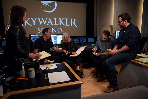 Five people sit in a circle in a recording studio discussing papers with a large logo behind them.